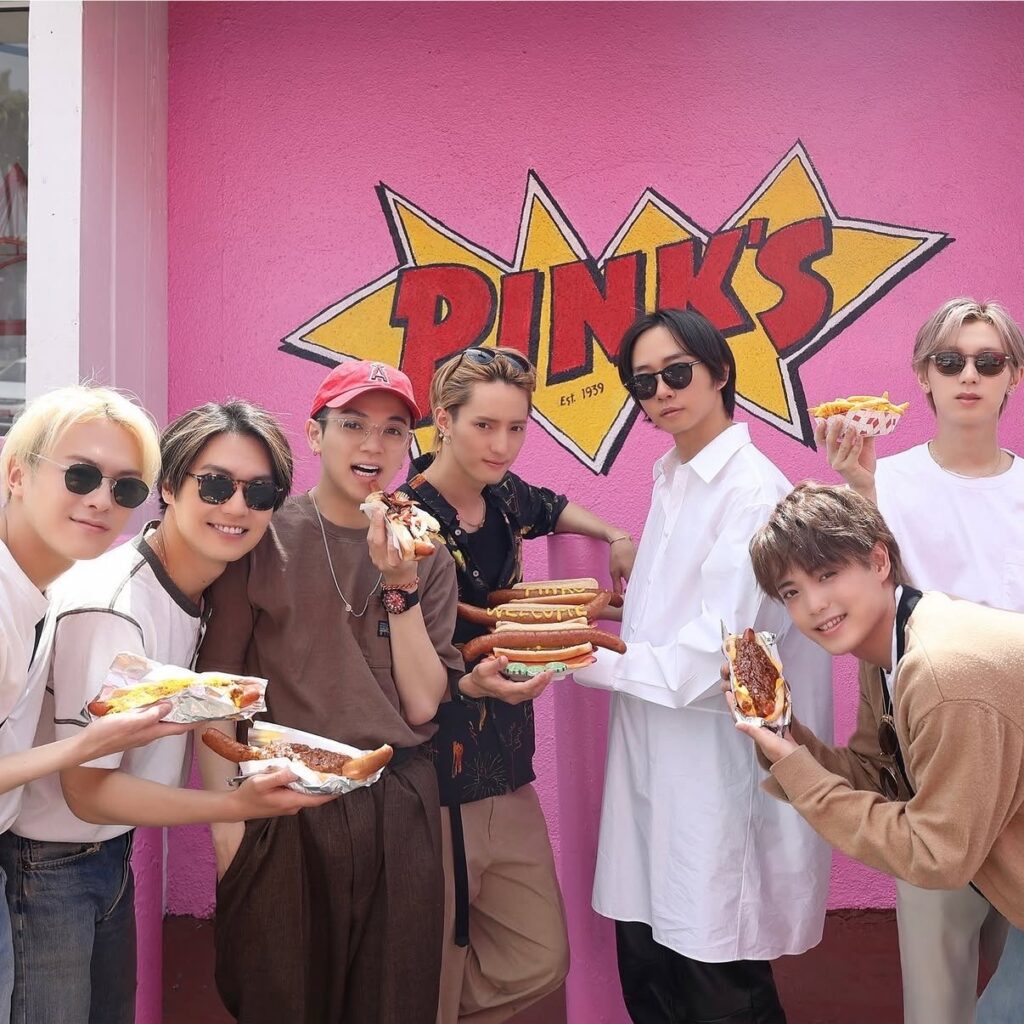 ピンクスホットドッグスの看板前でホットドッグを持つTravis Japanのメンバー7人